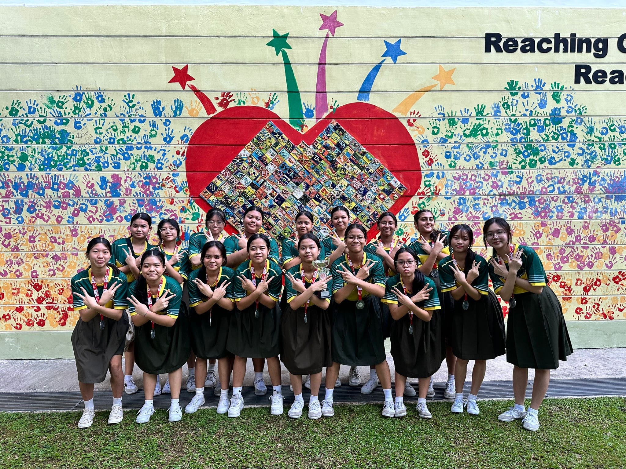 A school girls' floorball team posing together with a championship trophy in front of a vibrant wall mural featuring a large red heart and colorful handprints. The girls are wearing school uniforms and holding a uniform crossed-arm pose.